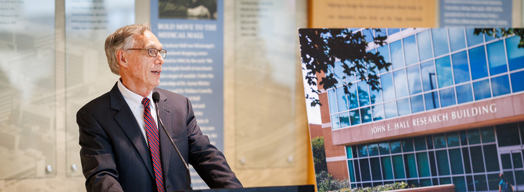 Dr. John Hall speaks at the podium during his retirement reception as an enlarged image of the newly-named John E. Hall Research Building is on a mounted poster to his left.
